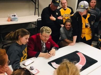 A man in a red jacket performs a card trick for a group of people in yellow uniforms gathered around a table.