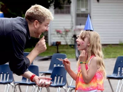 Man and girl with party hat face each other, both smiling with open mouths, outdoors near empty chairs.