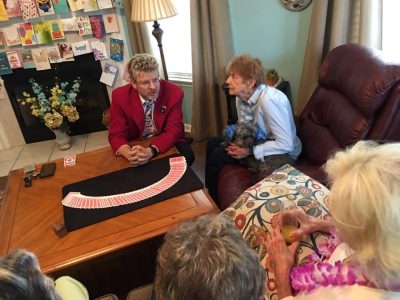 A man in a red jacket performs a card trick for elderly women seated around a table in a living room.