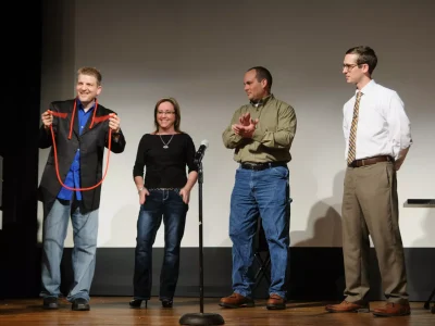 Four adults stand on stage, one holding a red rope, with a microphone in front of them and a table to the right.